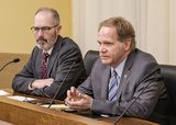 Tom Whaley sits at the testifiers' table and speaks to someone out of view. Representative Mahoney sits next to him.